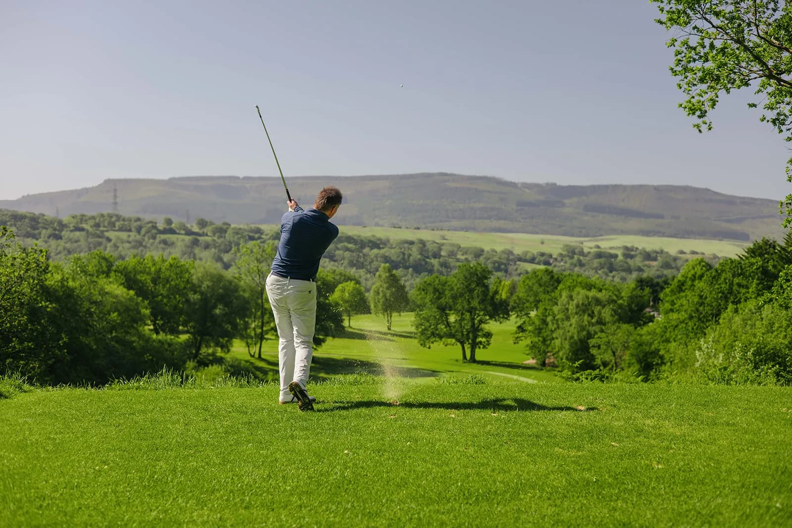 Stuart Wilson following through on a tee shot at Glynneath Golf Club, looking out over a wooded valley to the hills beyond